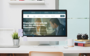 Un écran dordinateur affiche un site Web en français sur des services dassurance innovants. Le bureau est équipé de notes autocollantes, dune plante en pot, de livres, dun appareil photo et dun carnet de notes. Larrière-plan comprend une fenêtre avec des stores.