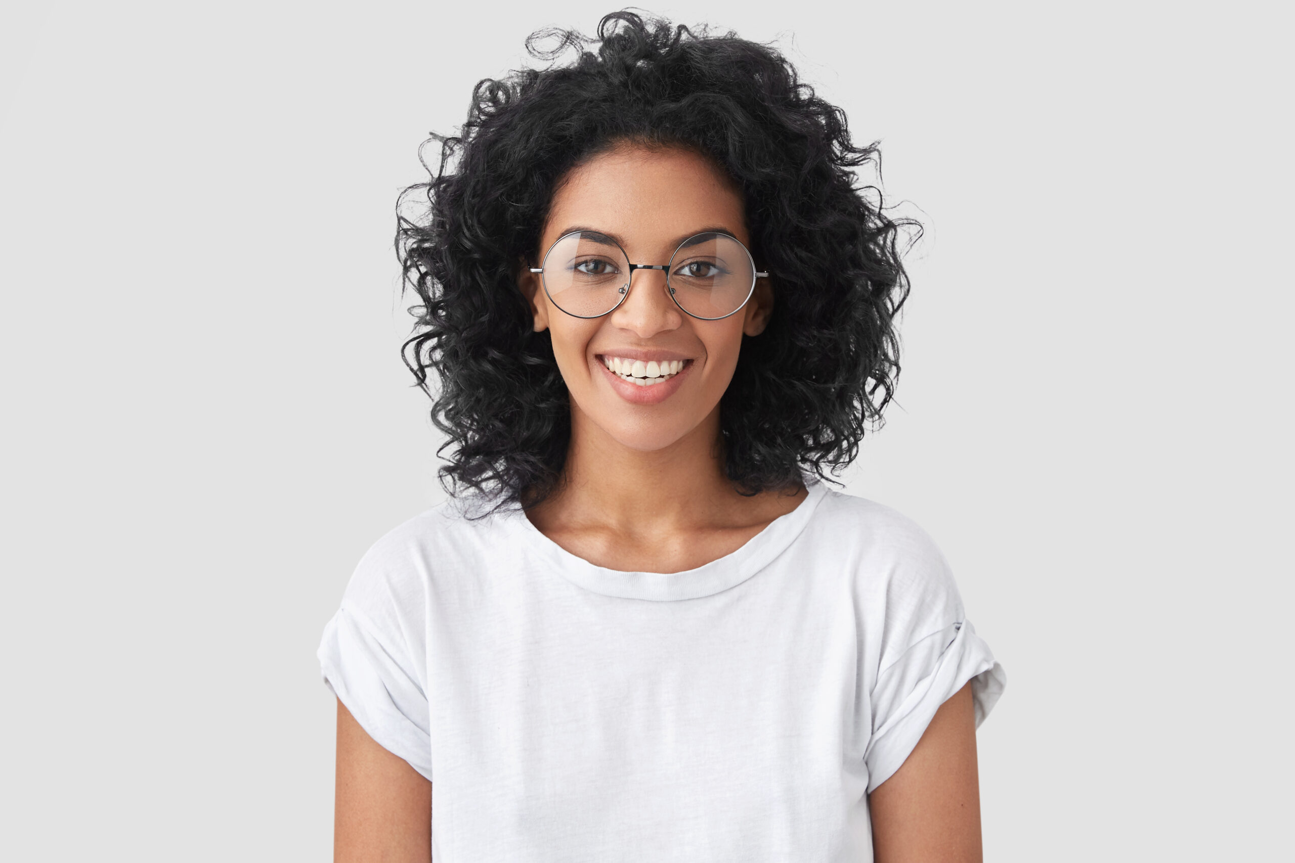 beautiful smiling african american female with crisp hair broad smile shows white teeth wears casual t shirt spectacles stands wall rejoices having day off woman journalist indoor scaled